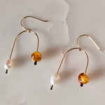 Load image into Gallery viewer, Pair of asymmetric arch gold fill earrings with gold glass beads, and pearls on a textured beige background.

